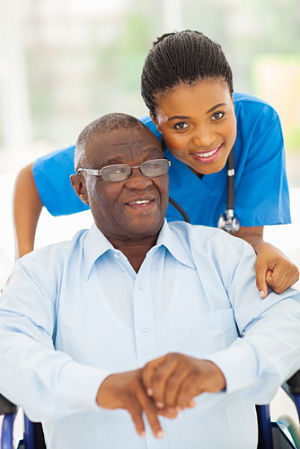 A female Nurse is standing behind an older male patient who is sitting down in a wheelchair. They are both smiling